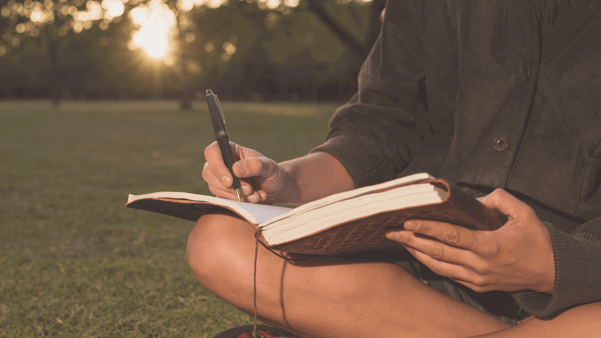 Person writing in a journal while sitting on the grass at sunset